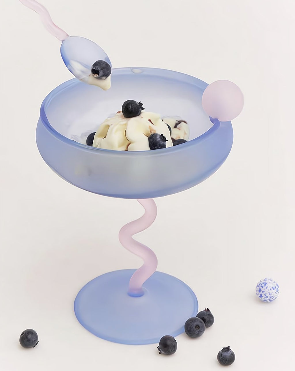 Blue dessert dish with a pink stand, topped with ice cream and blueberries, on a light gray background.